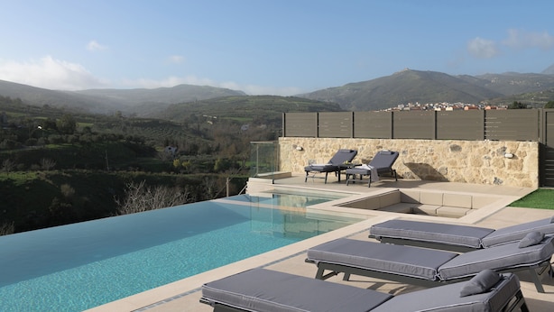 Piscina com espreguiçadeiras, parede de pedra e vista para colinas ao longe.
