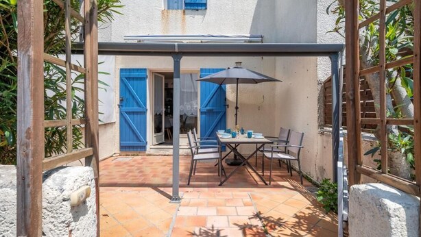 A covered patio with a table and chairs, blue shutters, and a white building in the background. - La Canotière - Close to the beach (Saint-Georges-d'Oléron)