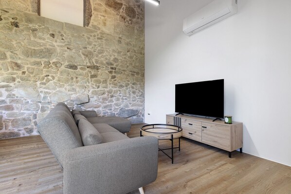 A modern living room with a stone wall, a sectional sofa, a TV on a wooden cabinet, and a round table. - Apartment 'T2 Familiar Ua01' with Shared Pool, Wi-Fi and Air Conditioning (Palmeira)