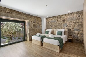 A bedroom with stone walls, two beds with green bedding, a small wooden nightstand, and a large window with a view of trees. - Apartment 'T2 Familiar Ua01' with Shared Pool, Wi-Fi and Air Conditioning (Palmeira)