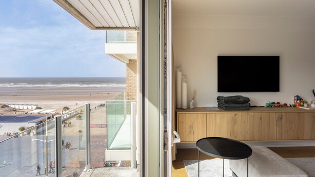 Ein modernes Wohnzimmer mit großem Fernseher, einem Balkon mit Glasgeländer und Blick auf den Strand.