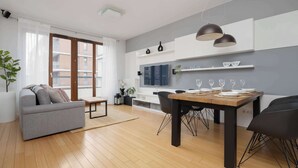 Ein modernes Wohnzimmer mit einer Couch, einem Esstisch und Stühlen. Es gibt eine Küchenzeile mit Fernseher und Regalen.