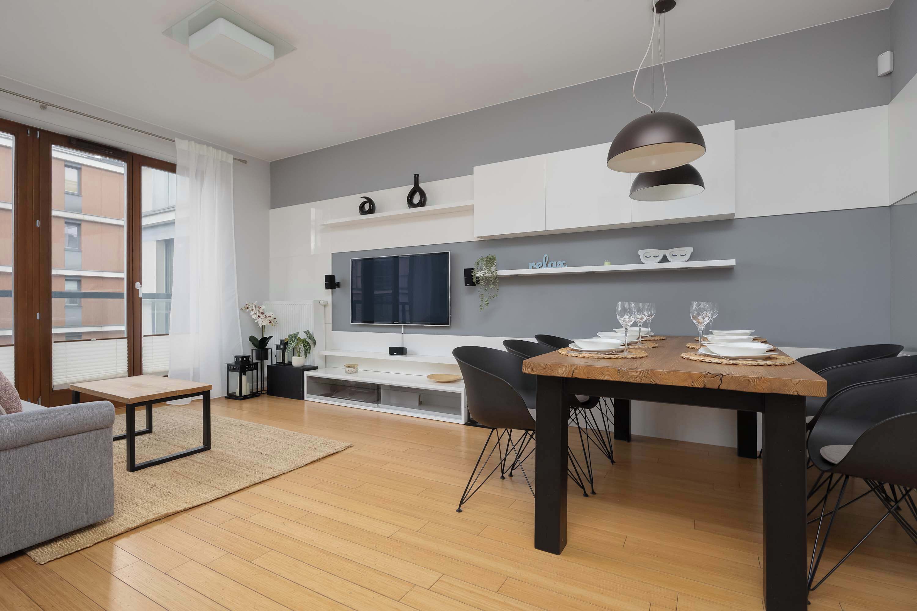 A modern living room with a wooden dining table, black chairs, a grey sofa, and a flat-screen TV mounted on the wall.
