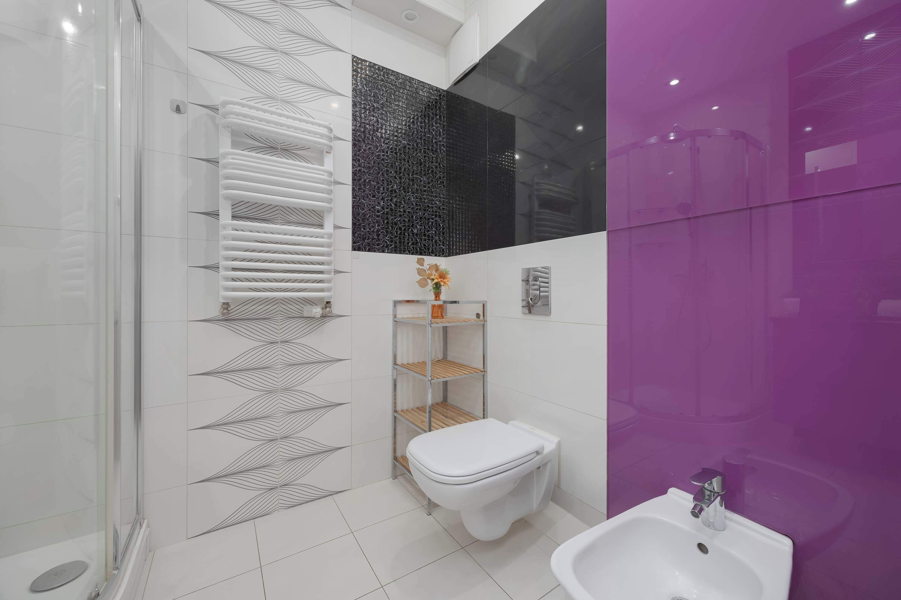 Un baño moderno con cabina de ducha de vidrio, paredes de azulejos blancos, inodoro blanco, lavamanos blanco y una pared de acento morada.