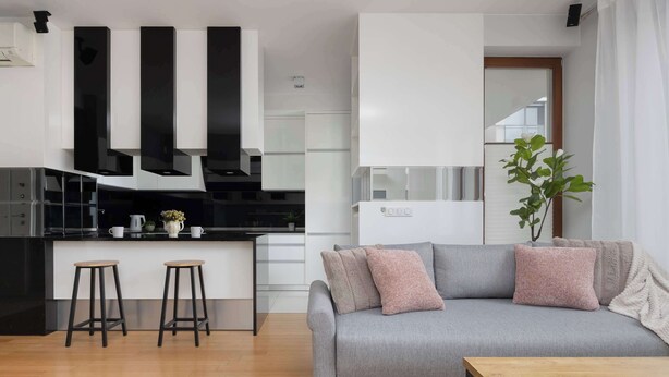 Ein modernes Wohnzimmer mit grauem Sofa, zwei Holzschemeln, einem Holztisch und einer Küchenzeile mit weißen Schränken und schwarzen Akzenten.