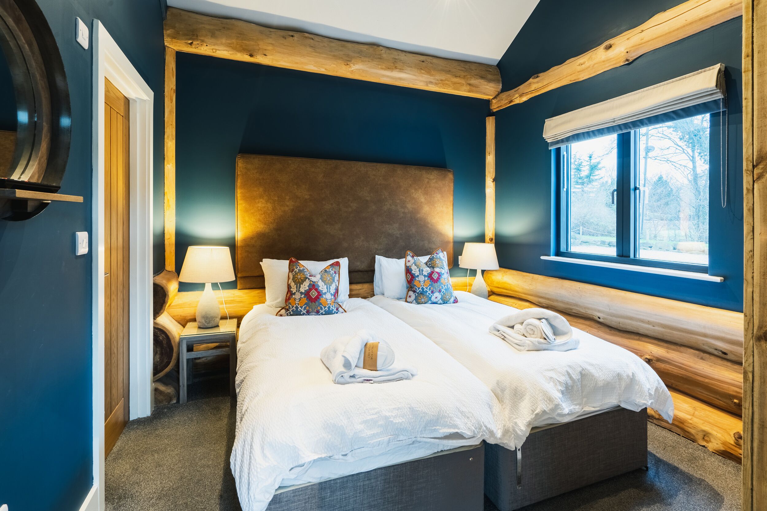 Cama doble con sábanas blancas, dos mesitas de noche con lámparas, cabecera de madera y ventana con vistas a árboles.
