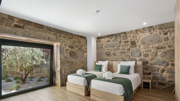 A bedroom with stone walls, two beds with green bedding, a small wooden nightstand, and a large window with a view of trees. - Apartment 'T2 Familiar Ua02' with Shared Pool, Wi-Fi and Air Conditioning (Palmeira)