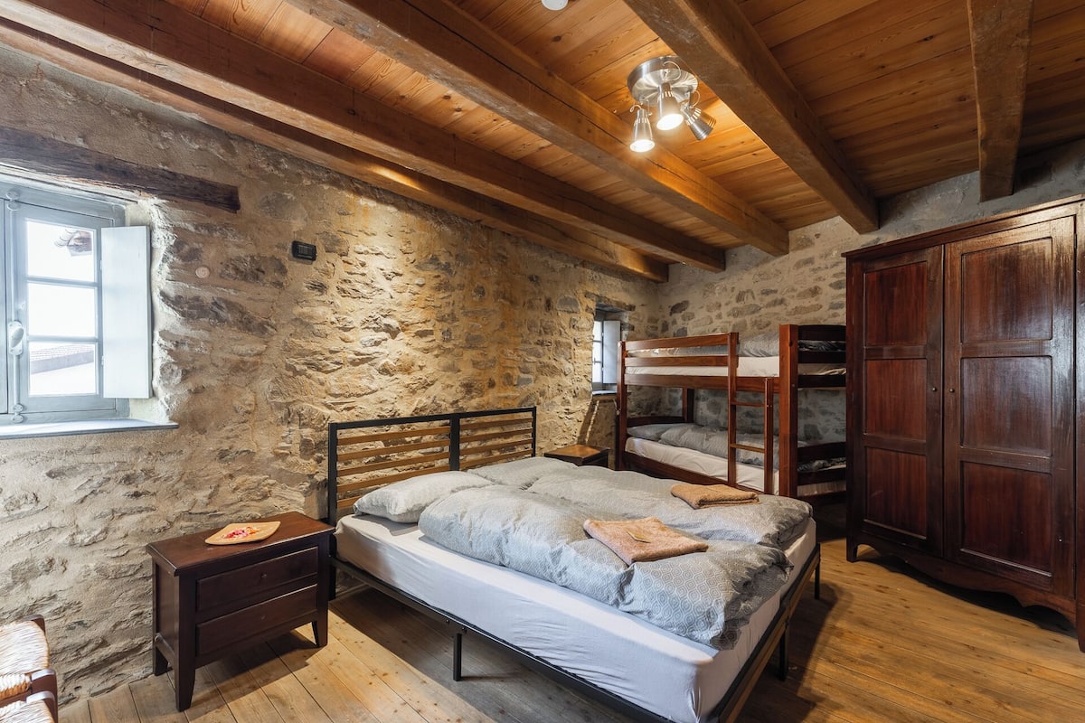 Un dormitorio rústico con pared de piedra, vigas de madera en el techo, una litera, un armario de madera y una mesita de noche con lámpara.