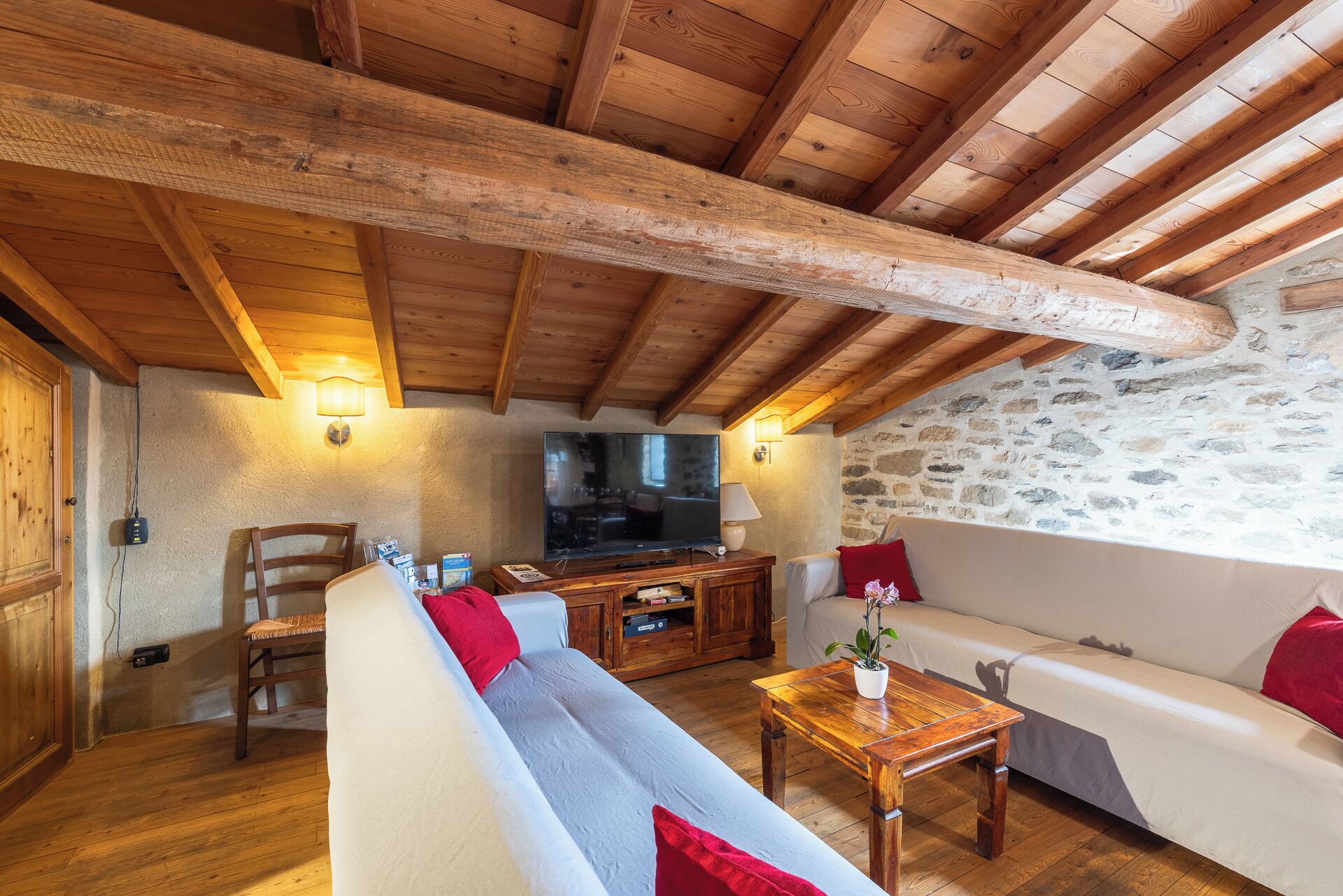A rustic wooden interior with a stone wall, wooden ceiling beams, a sofa, a television, and a wooden table.