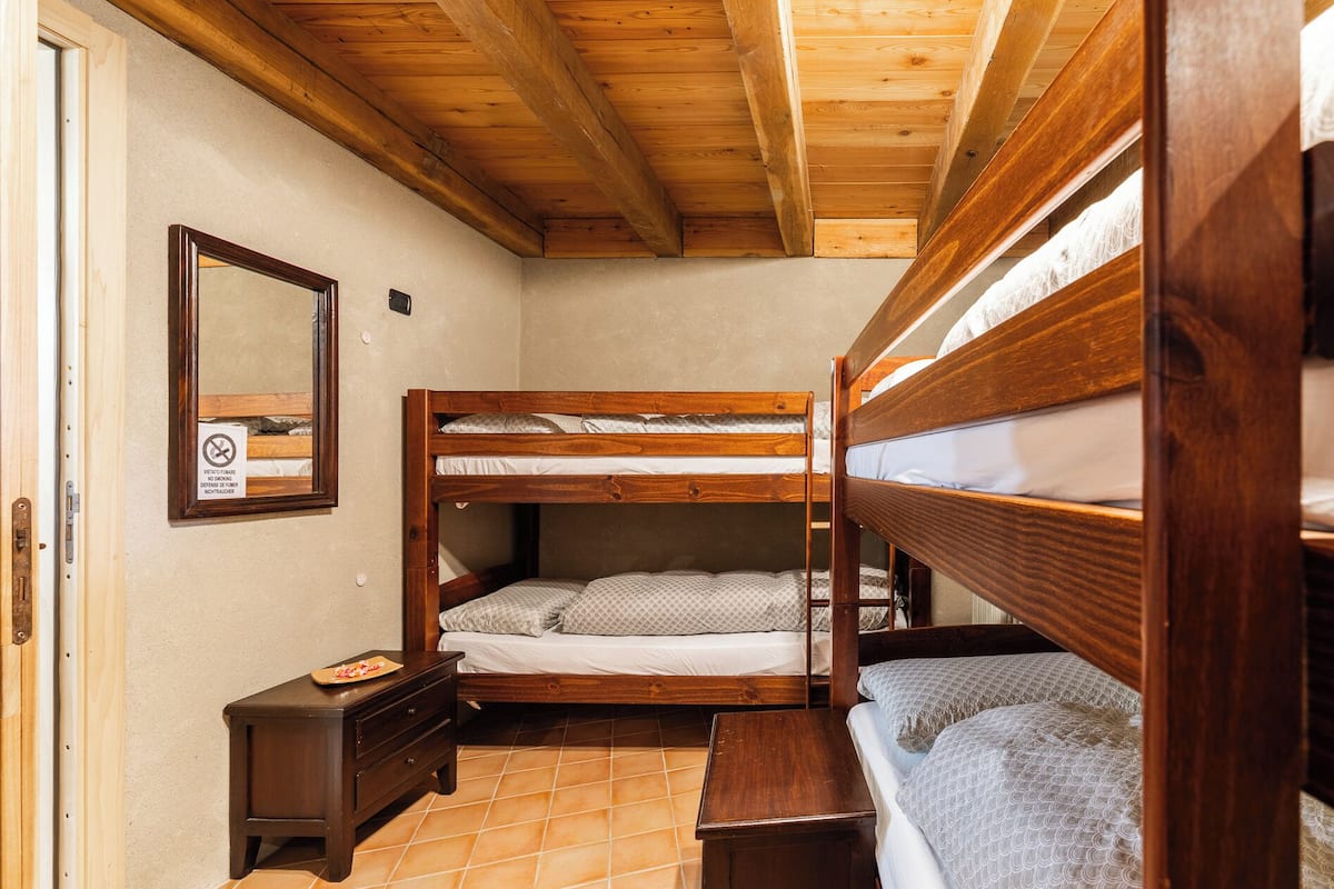 Pequeña habitación con literas de madera, suelo de baldosas, una mesita de noche de madera y un espejo en la pared.
