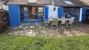 Una casa rústica con techo de piedra, contraventanas azules y un comedor con mobiliario exterior.
