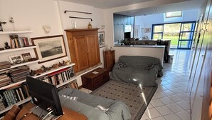 Amplia sala de estar con una cama grande, un escritorio con computadora, una estantería y vistas al exterior a través de grandes ventanales.