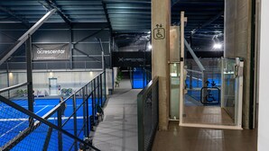 Instalación deportiva interior con cancha de tenis, escalera y ascensor de vidrio.