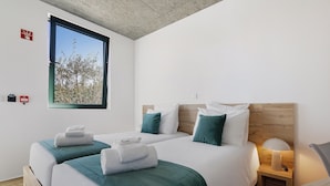 Habitación de hotel con dos camas, ventana con vista a árboles, mesita de noche con lámpara y cabecera de madera.