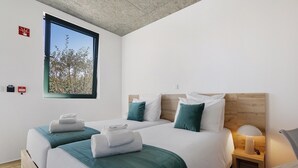 Ein Hotelzimmer mit zwei Betten, einem Fenster mit Blick auf Bäume, einem Nachttisch mit Lampe und einem hölzernen Kopfteil.