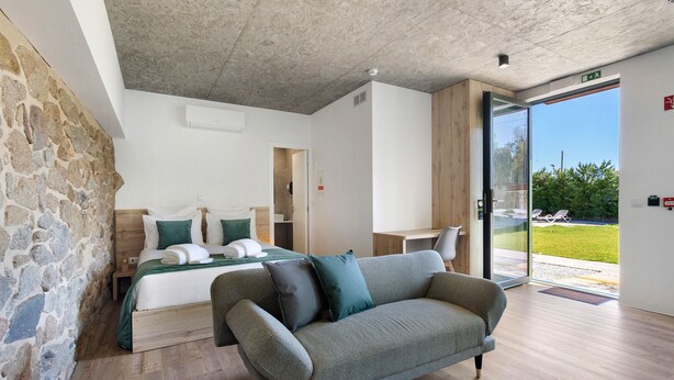 Une chambre d’hôtel moderne avec un mur en pierre, un lit avec une literie verte, un canapé gris et une grande porte-fenêtre coulissante donnant sur un espace extérieur.
