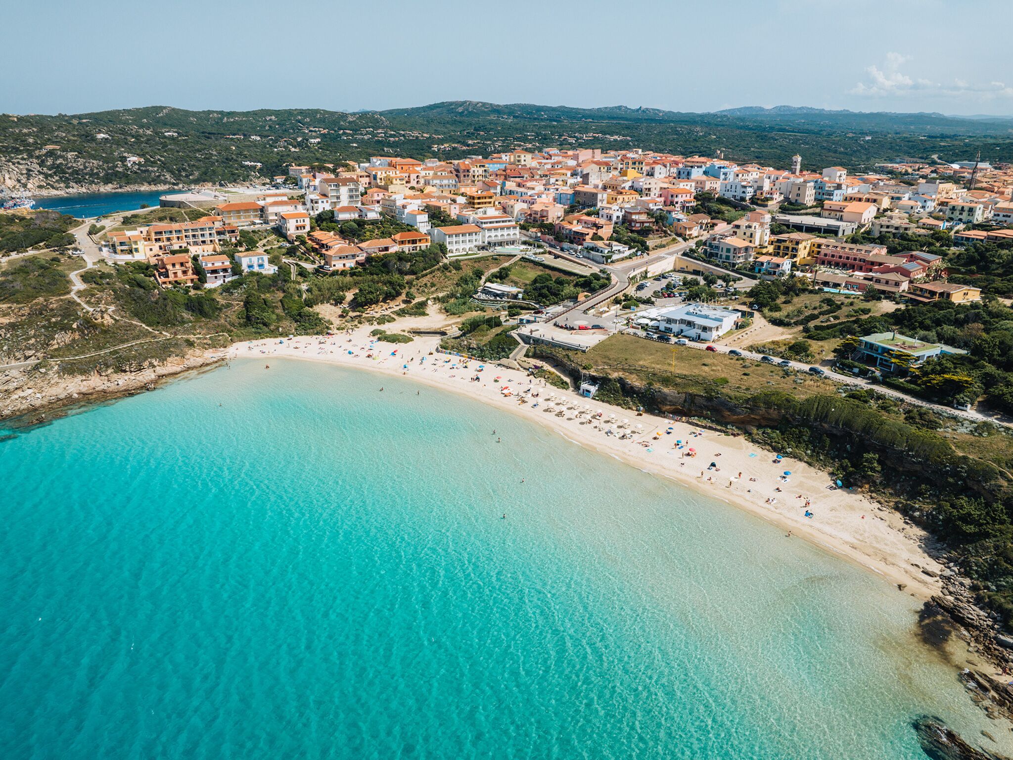 Vista aérea de un pueblo costero con playa arenosa, edificios y mar azul cristalino.