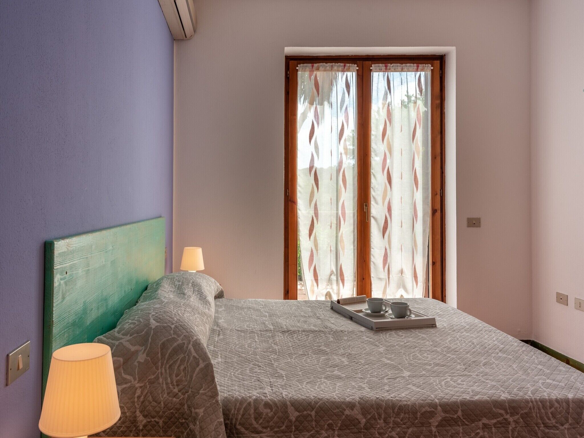 A bedroom with a bed, a bedside lamp, a window with curtains, and a tray with cups and a saucer on the bed.