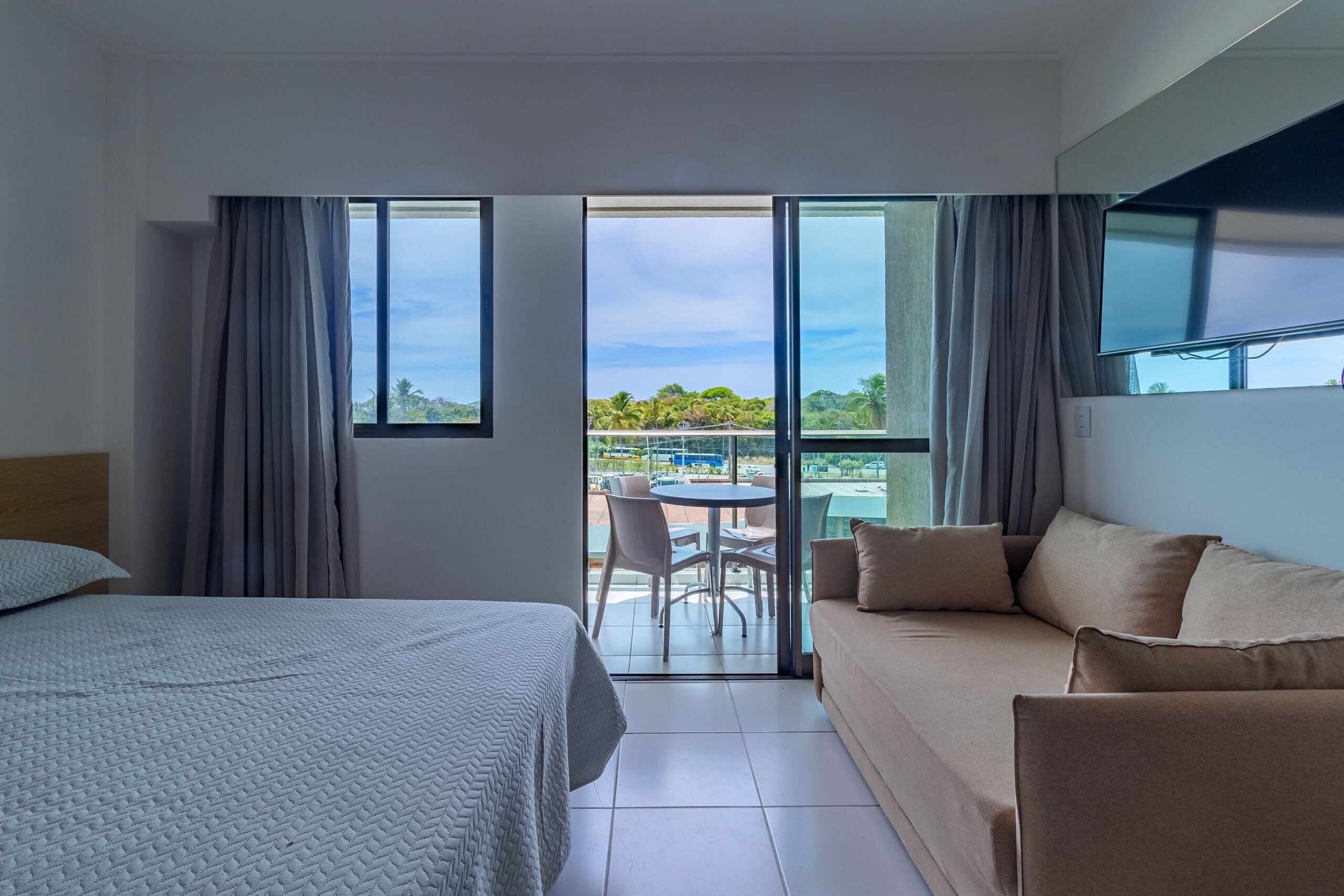 Quarto de hotel com cama, sofá, varanda com mesa e cadeiras, e vista para o verde.
