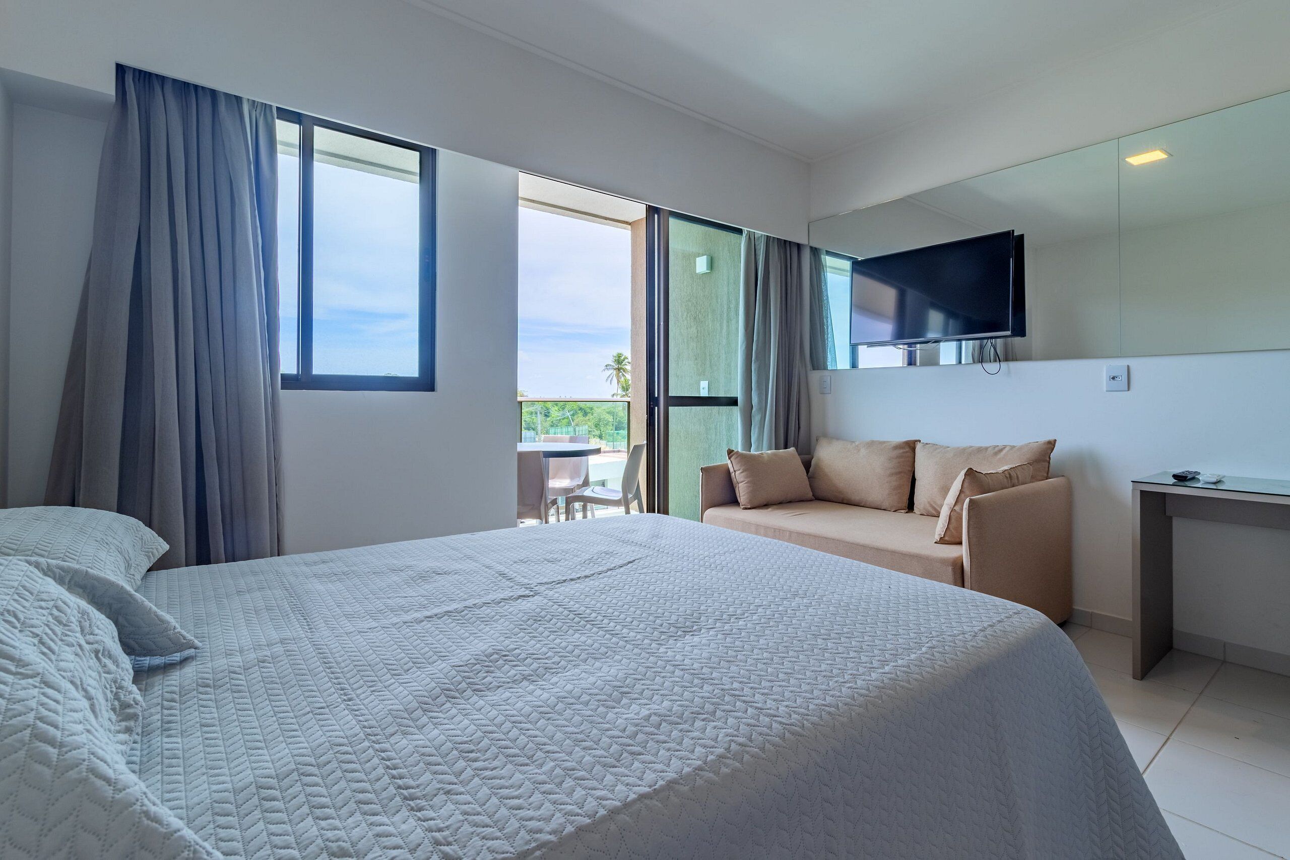 Quarto de hotel com uma cama grande, um sofá, uma TV na parede e uma varanda com vista.