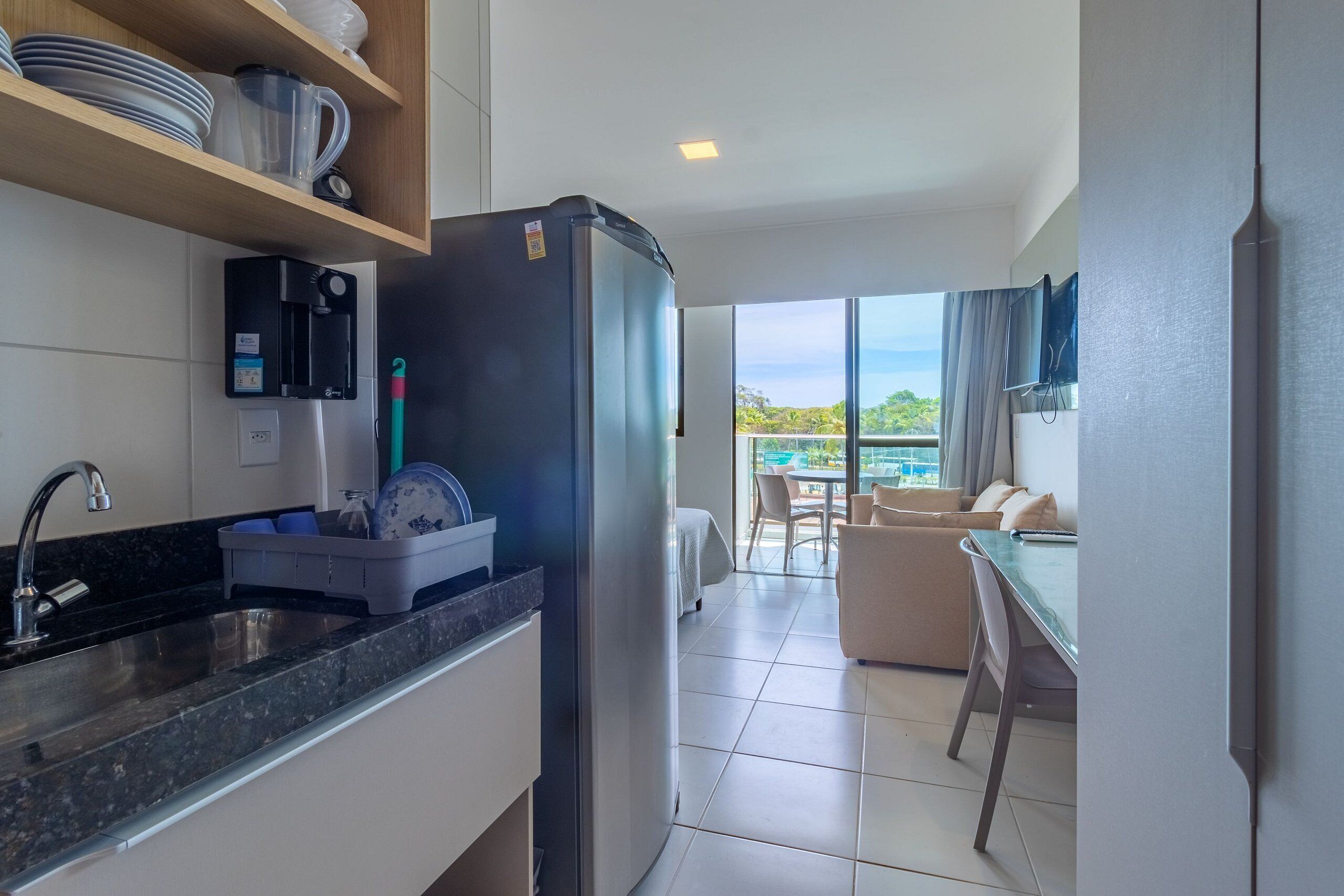 A modern kitchen with a refrigerator, sink, and dish rack. There is a dining area with a table and chairs, and a balcony with a view.