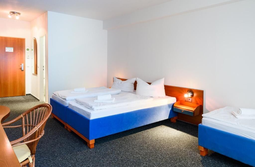 Une chambre d’hôtel avec un lit simple, un petit lit, une chaise, un bureau et une table de chevet avec une lampe.