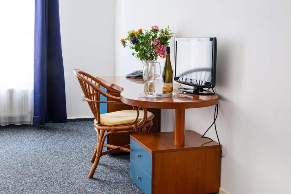 Quarto de hotel com escrivaninha de madeira, uma cadeira, televisão, um vaso com flores e uma garrafa de vinho.