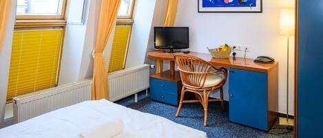 Een hotelkamer met een bed, een bureau met een computer, een stoel, een raam met luxaflex en een schilderij aan de muur.
