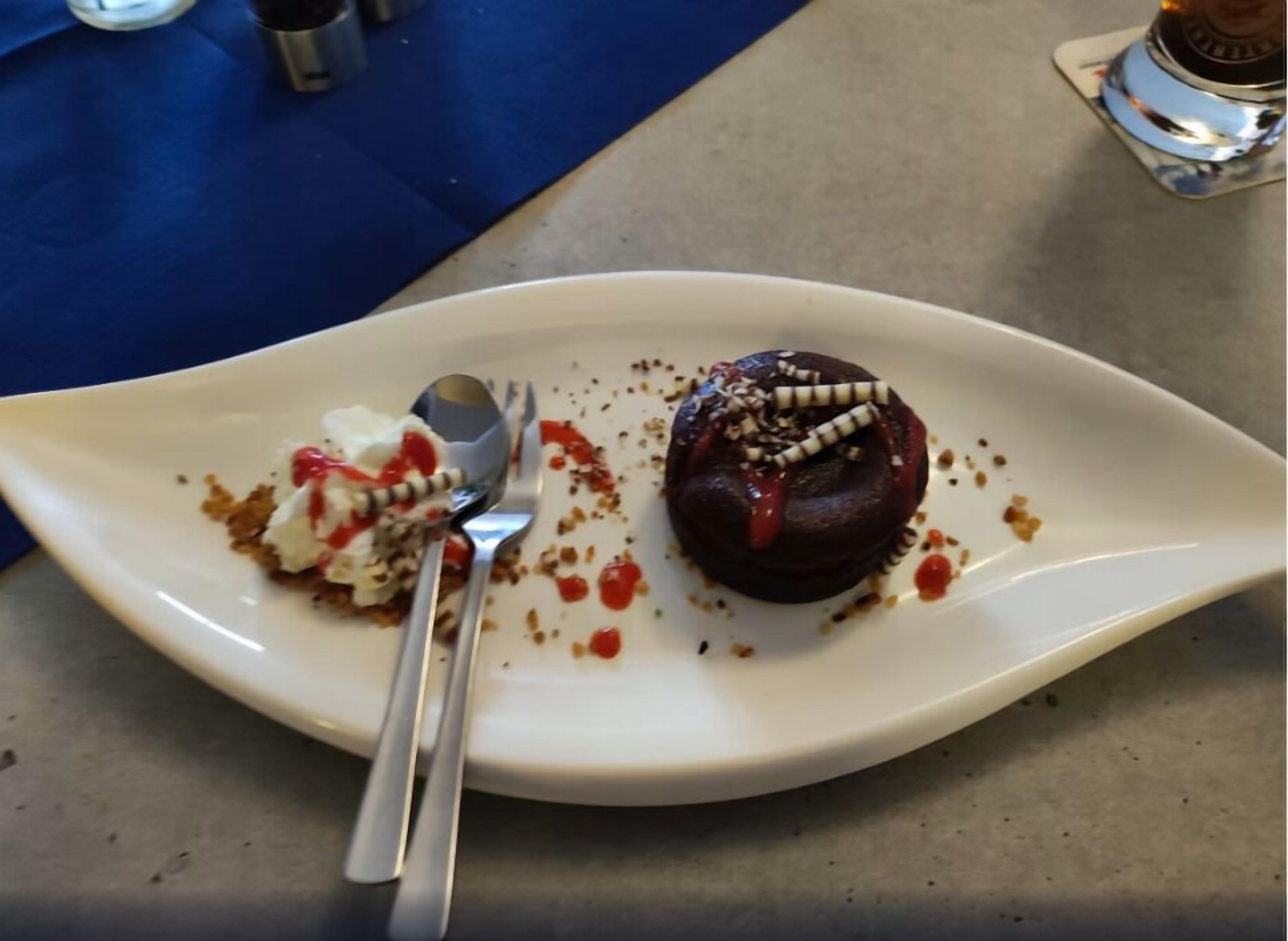 Un postre en un plato blanco, con una cuchara, migas y un mantelino azul al fondo.