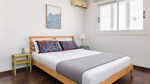Ein Schlafzimmer mit einem hölzernen Bett, gemusterten Kissen, einer Nachttischlampe, einem kleinen Tisch, einem gerahmten Bild und einem Fenster mit Jalousien.