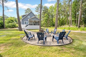A house with a porch, a fire pit surrounded by chairs, and a forested area. - Relax Inn by Seaside Vacations & Sales (Chincoteague Island)