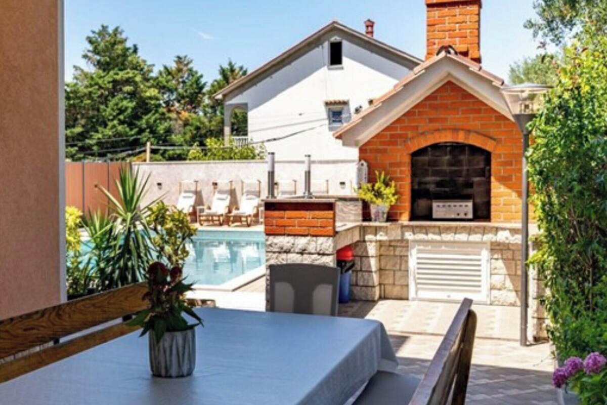A poolside area with a table, a brick oven, and a white building in the background.