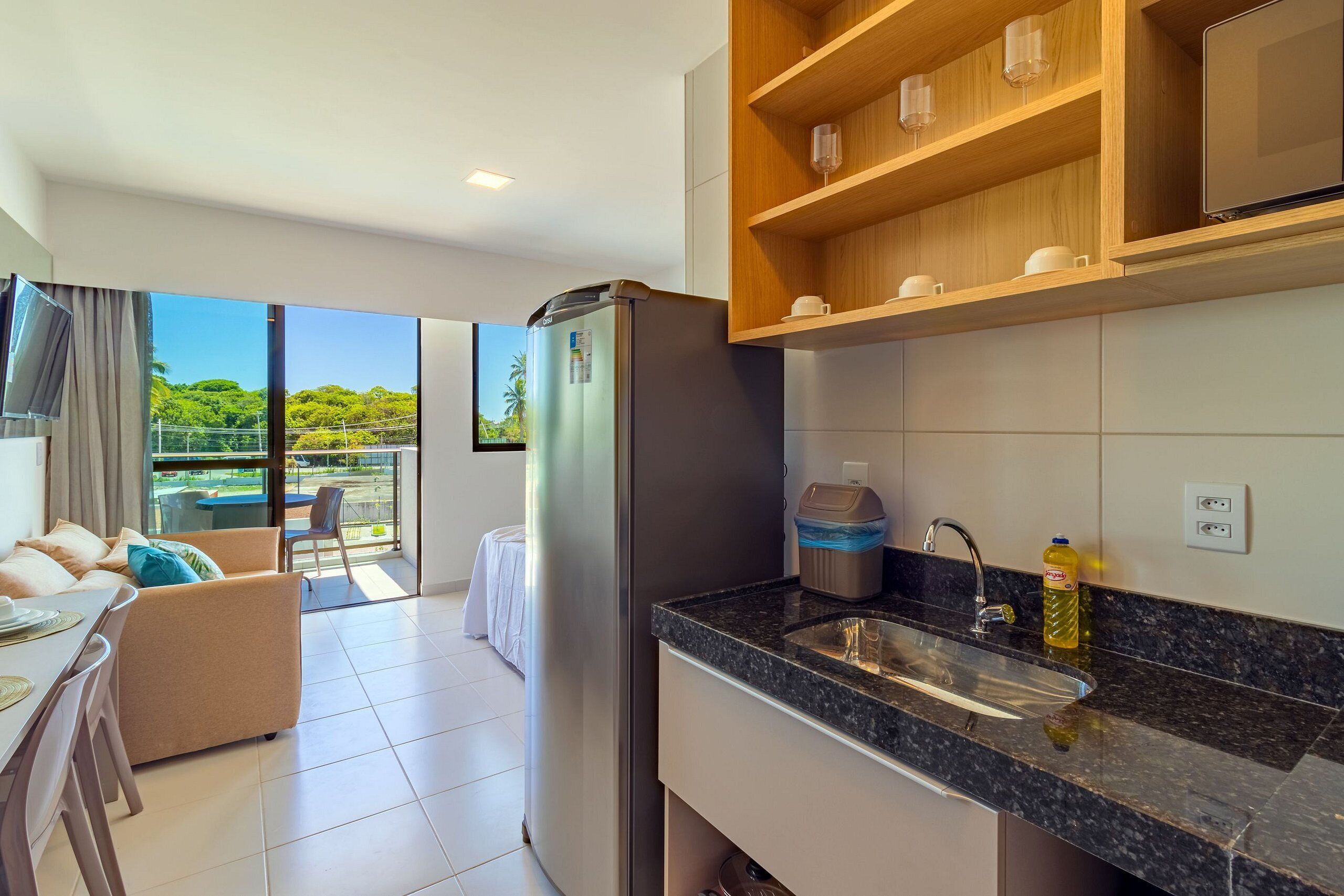 Una cocina moderna con refrigerador, pileta y estantes de madera. Hay un comedor con mesa y sillas, y un balcón con vista a espacios verdes.