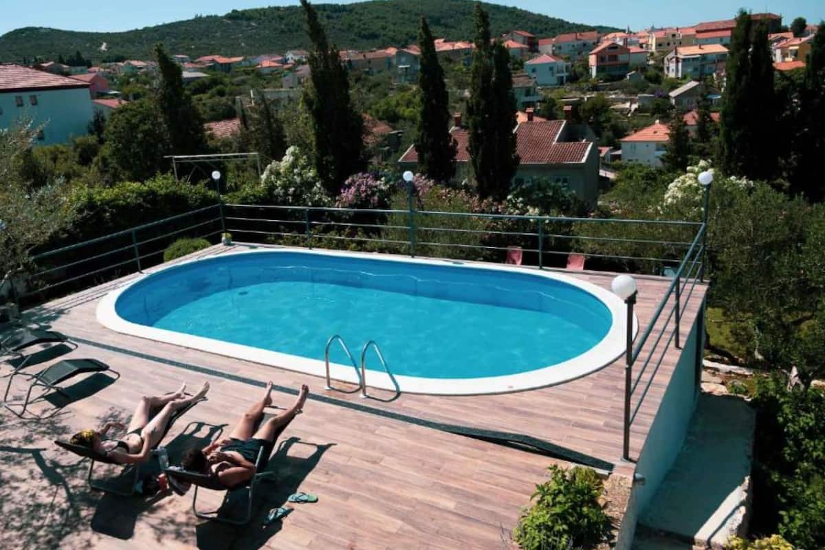 A swimming pool with a wooden deck, two lounge chairs, and a view of a hillside town.