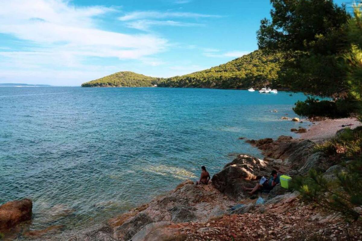 A rocky shoreline with clear blue water, a distant hill with trees, and a boat in the background.