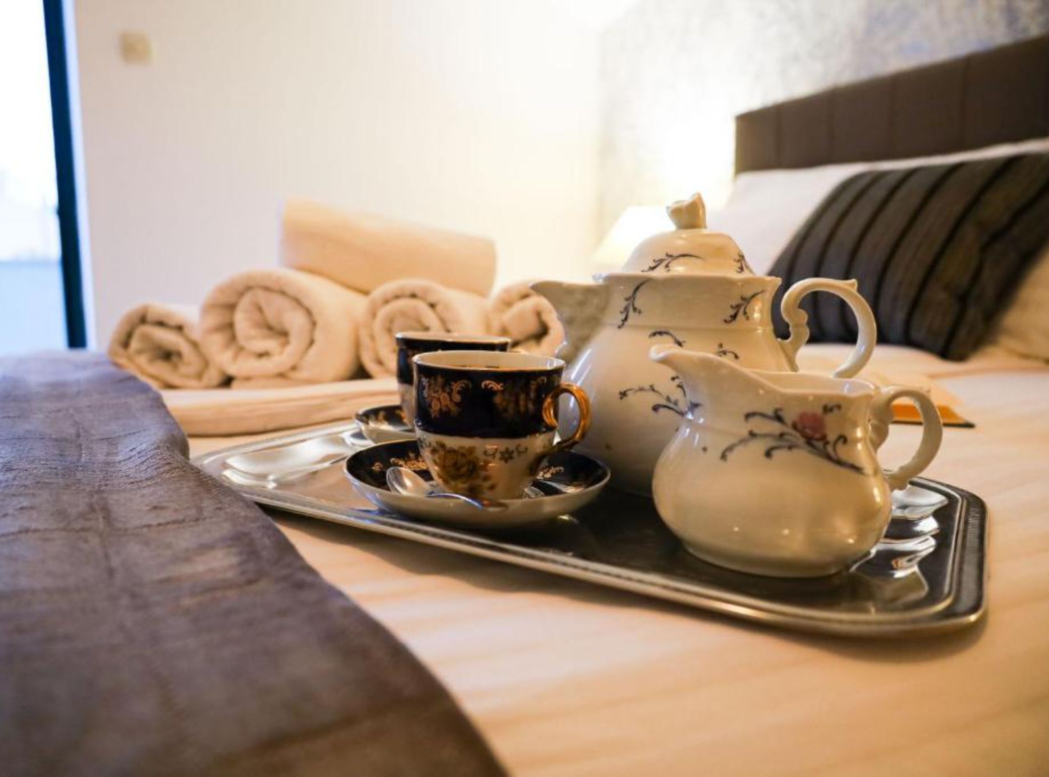 Una bandeja con un calentador de agua, una taza y toallas enrolladas sobre una cama.