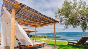 A wooden pergola with a white canopy, a pool, and lounge chairs on a grassy deck with a view of the sea and hills.