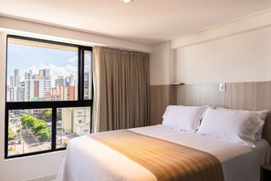A hotel room with a large window, a bed with white linens, and a view of a cityscape. - Move #914 - Apartamento Comfort by Carpediem (João Pessoa)