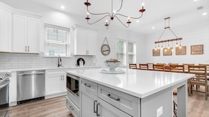 A modern kitchen with white cabinetry, a central island, stainless steel appliances, and a dining area with wooden chairs. - Latitude! New! 8 Bed! Beach! (Destin)