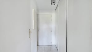 Ein schmaler Korridor mit weißen Wänden und Fliesenboden, der zu einer geschlossenen Tür führt.