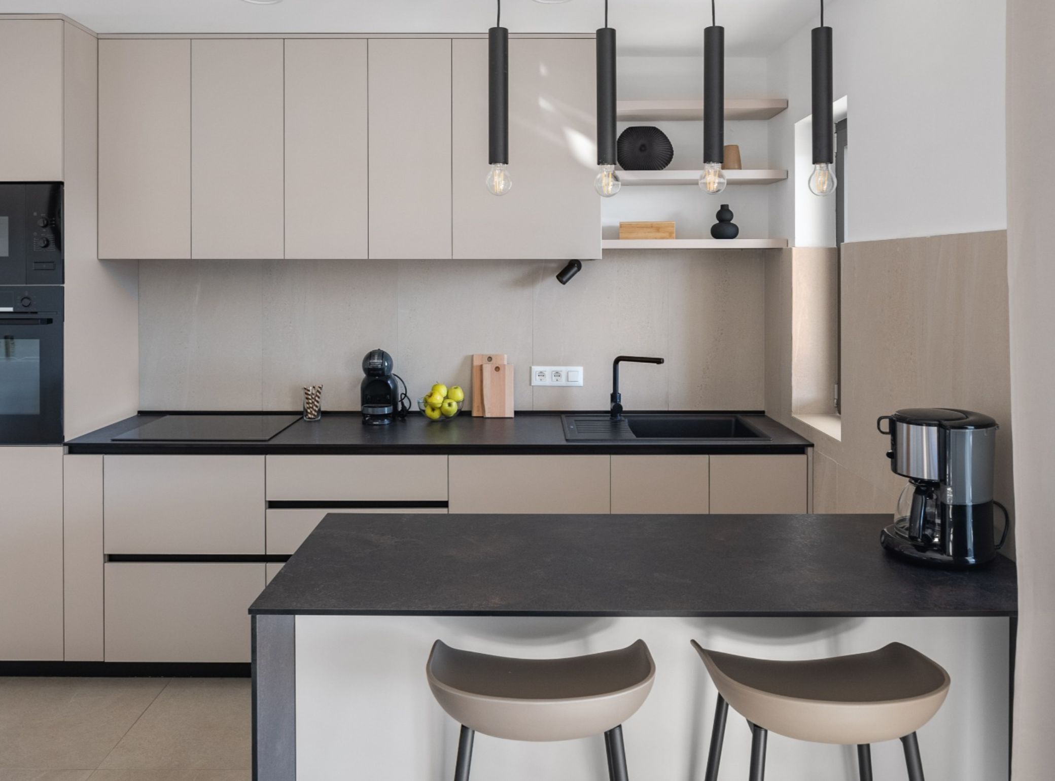 Una cocina moderna con encimera negra, dos taburetes de bar, una cafetera y un horno empotrado.