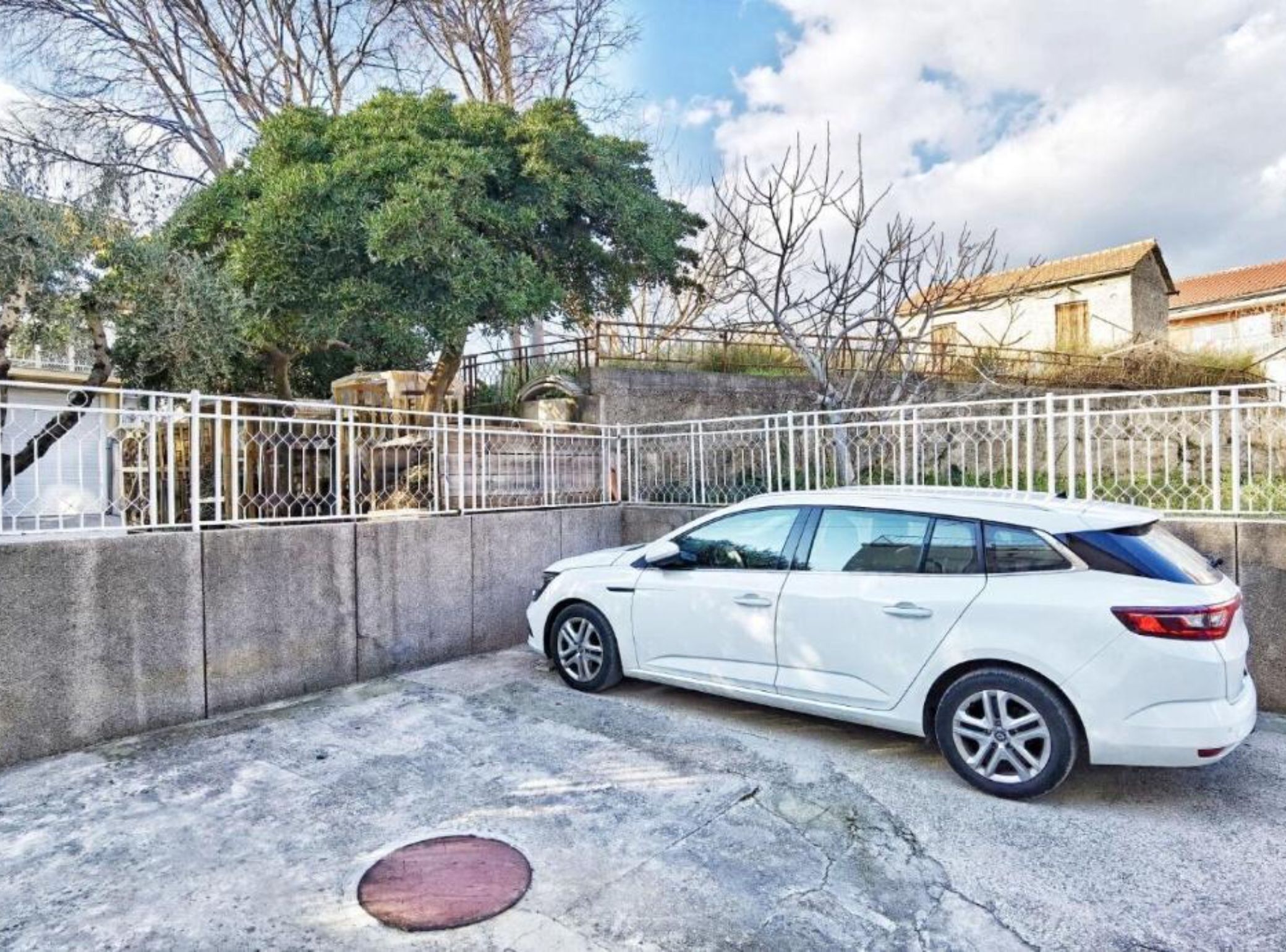 A white car parked in a driveway with a concrete wall and a metal fence.