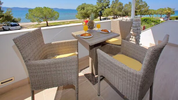 A balcony with wicker furniture, a table with breakfast items, and a view of the sea and trees. - Apartments Blue Horizon Retreat (Zadar)