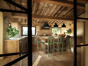 Une cuisine rustique avec des poutres en bois, un grand îlot central doté de chaises et une vue sur l’extérieur.