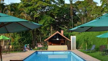 Una piscina con sillas de descanso y sombrillas, rodeada de vegetación y un edificio al fondo.