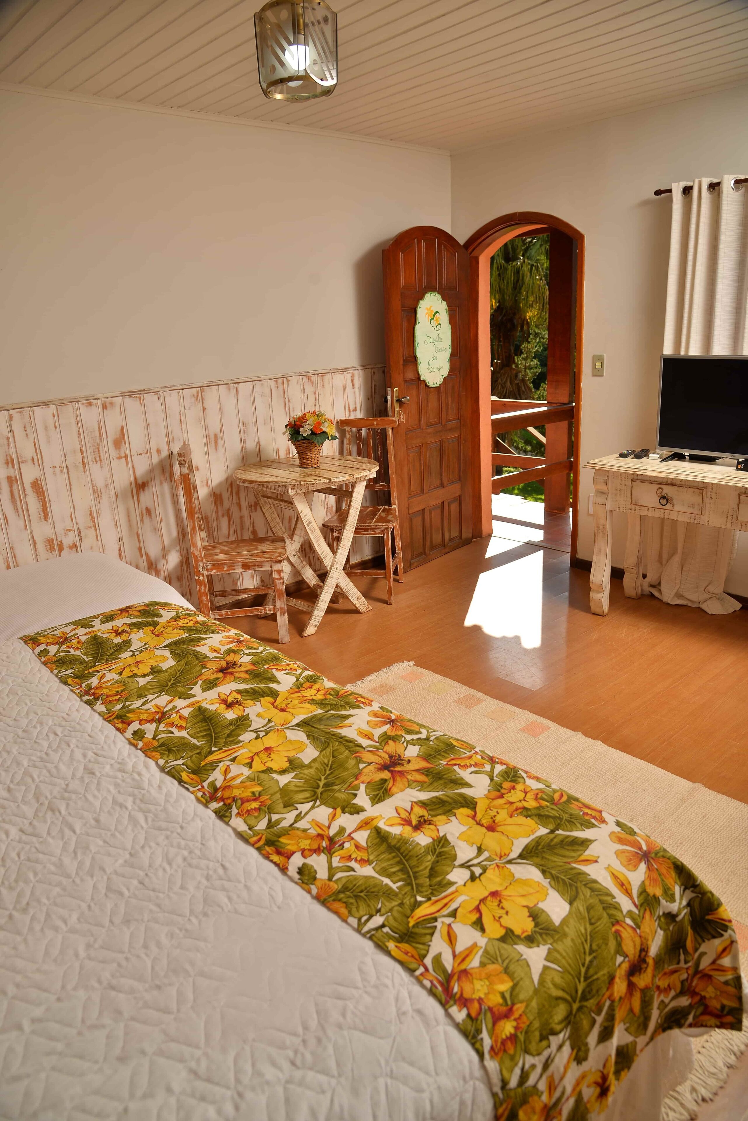 A bedroom with a bed, a wooden desk, a television, and a door leading to a balcony.