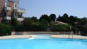 A swimming pool with a curved edge, surrounded by a low fence, and adjacent to a multi-story building with balconies. - Golf Apartment - Pornic 2 with Shared Pool, Balcony and Wi-Fi (Pornic)