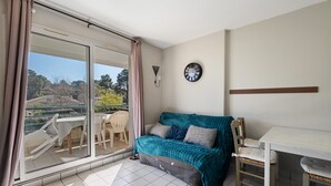 A room with a sliding glass door leading to a balcony, a teal sofa, a white table, and a chair. - Golf Apartment - Pornic 2 with Shared Pool, Balcony and Wi-Fi (Pornic)