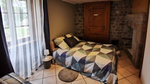 A bedroom with a bed, a wooden door, a stone wall, and a window with curtains. - Gîte du Platane, Sleeps 14 (Grazac)