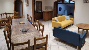 A dining area with a long table and chairs, a sofa, a small round table, and a kitchen area with cabinets and a clock. - Gîte du Platane, Sleeps 14 (Grazac)